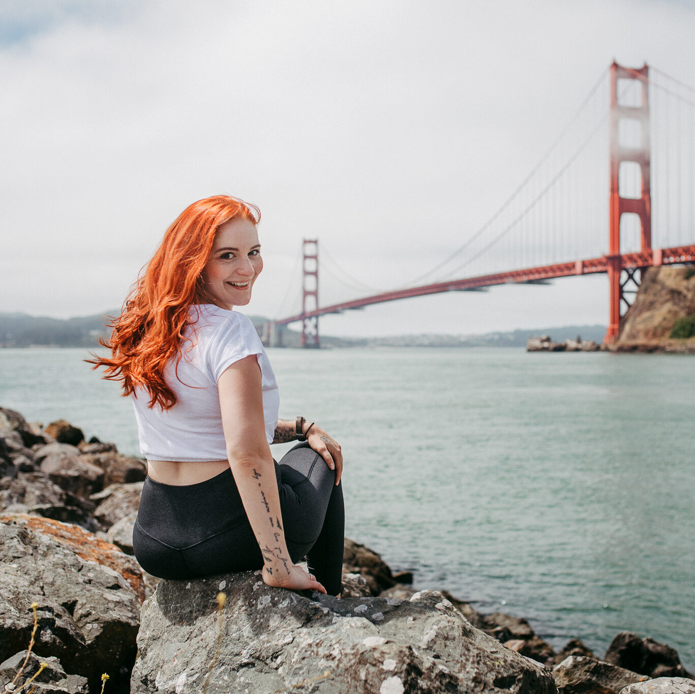 Fotografin Kim Kärger sitzt auf ein paar Steinen am Meer mit dem Ausblick auf die Golden Gate Bridge in San Francisco. Blickt nach hinten in die Kamera und lächelt.