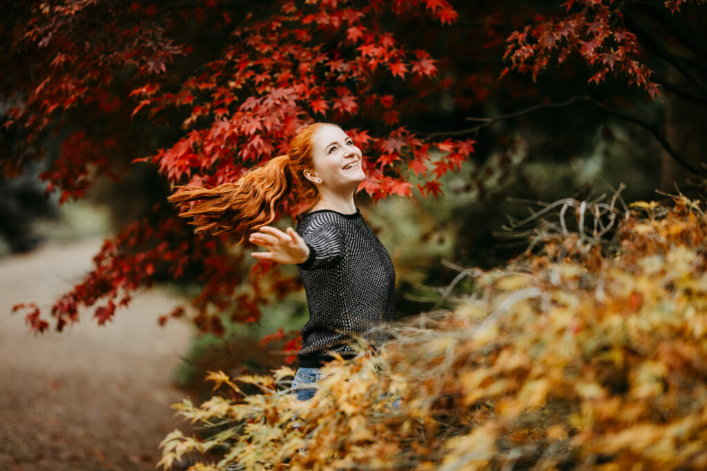 Fotografin Kim Kärger dreht sich im Kreis und lächelt in die Luft. Um sie herum sind viele bunte Herbstblätter zu sehen. Die Haare fliegen in der Bewegung.