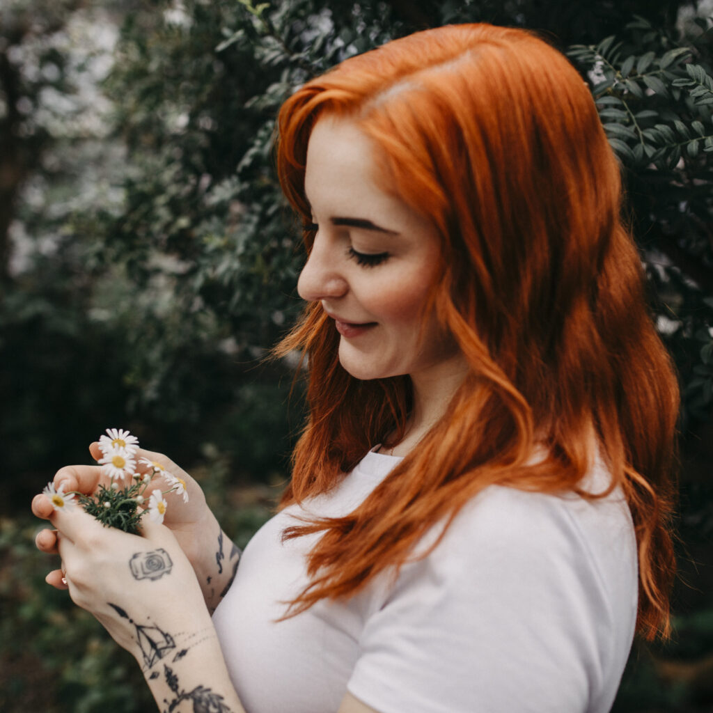 Detailbild von Kim Kärger die Gänseblümchen in den Händen hält. Auf der Hand ist eine kleine Kamera tätowiert.